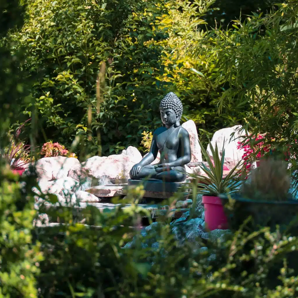 Statue de Bouddha méditant en pierre sombre, entourée de feuillage vert luxuriant et de rochers clairs dans un jardin zen paisible.