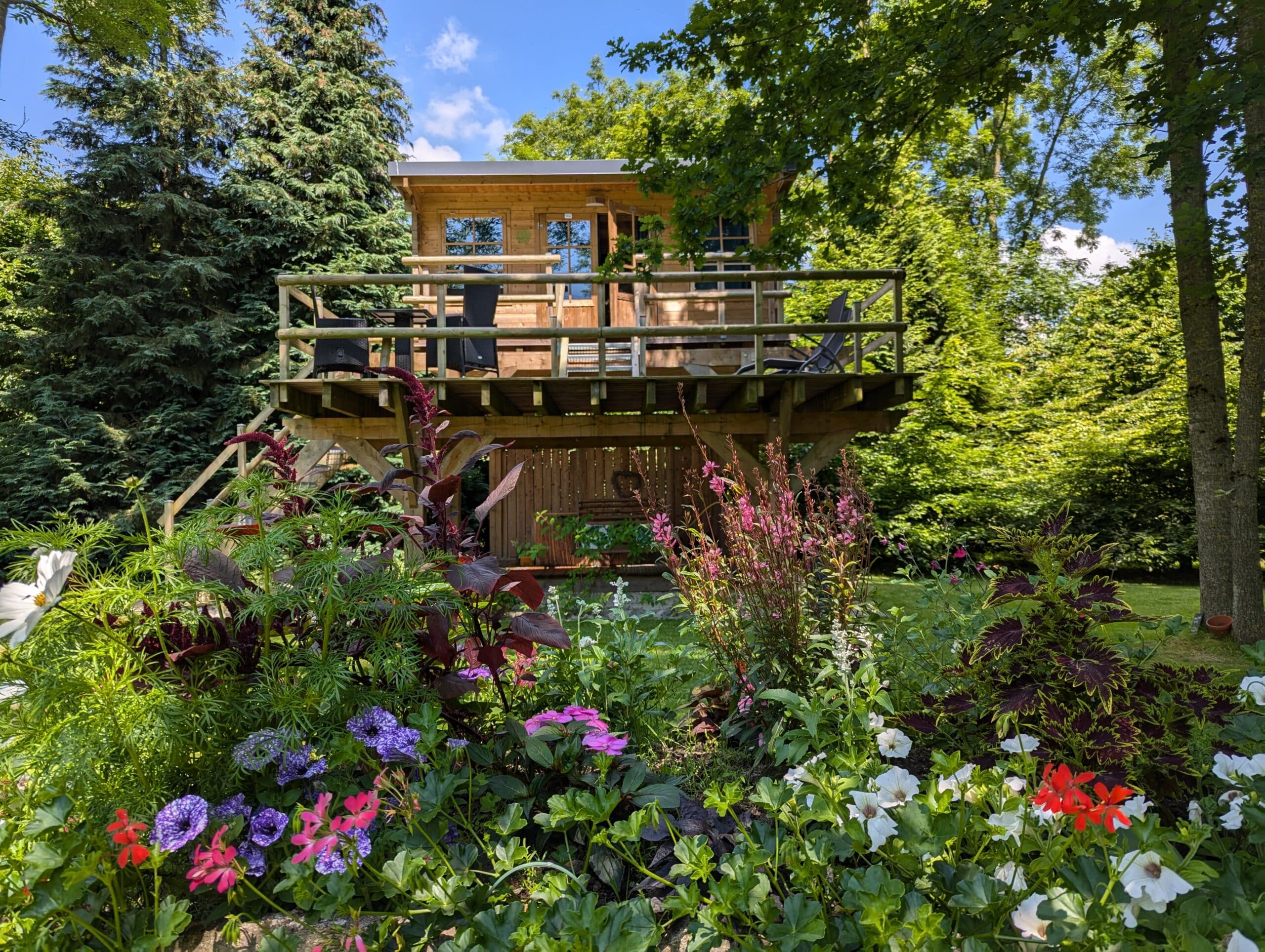 Invitante cabane en bois surélevée avec terrasse et salon de jardin, au milieu d'un luxuriant jardin fleuri sous un ciel bleu ensoleillé.