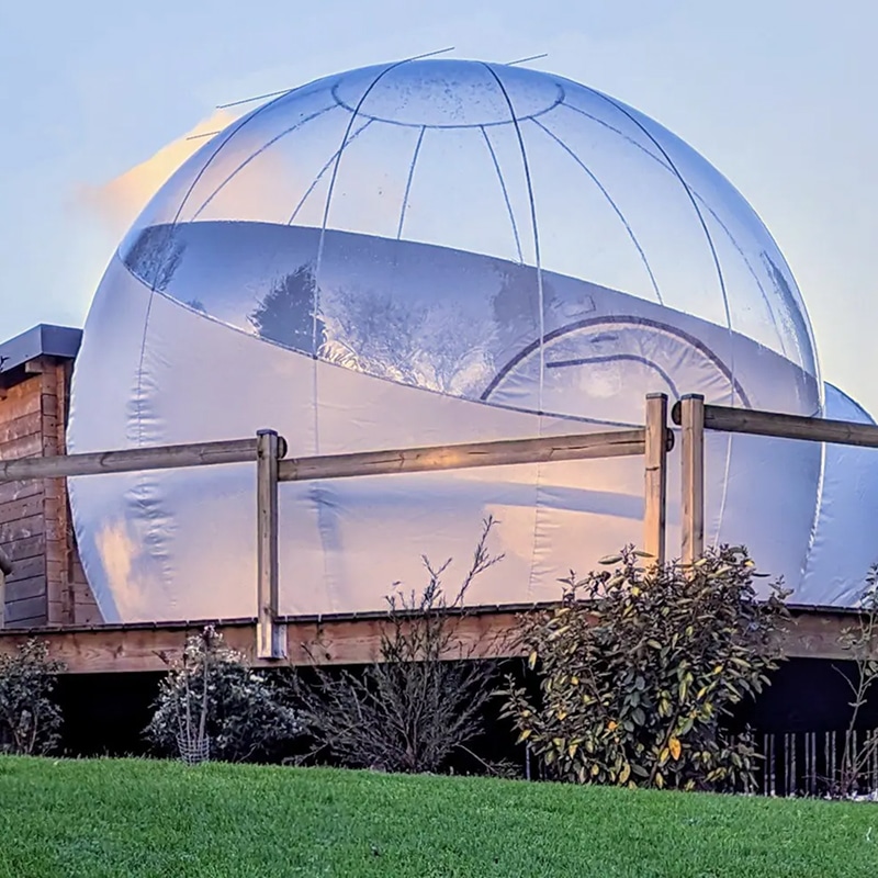 Grande bulle transparente avec base blanche sur terrasse en bois, entourée de verdure, sous ciel clair. Expérience insolite.