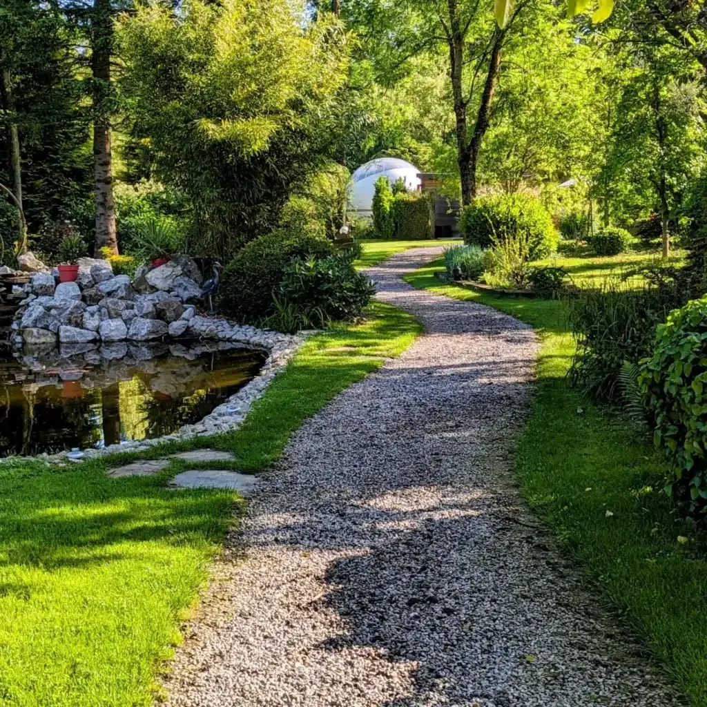 Chemin de gravier serpentant dans un jardin luxuriant, menant à un dôme blanc moderne, avec un étang rocheux à gauche.