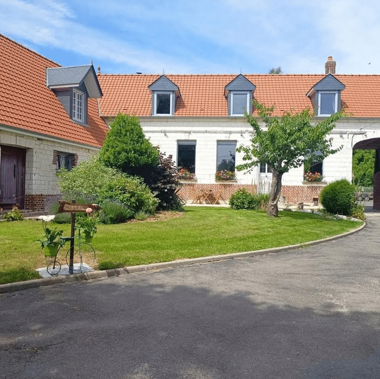 Charmante maison traditionnelle au toit tuile rouge et murs clairs, entourée d'un jardin verdoyant. Panneau "Heure & O'd'Champs", fenêtres fleuries.
